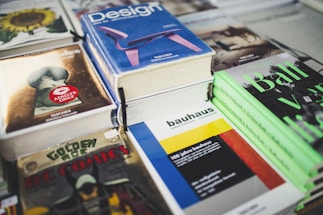A collection of books and magazines is arranged on a table, including titles such as 'Design', 'bauhaus', 'Golden Age DC Comics', and a book with an 'Adults Only' label. The covers display bold colors and graphics, featuring a variety of subjects from comics to architecture.