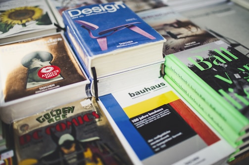 A collection of books and magazines is arranged on a table, including titles such as 'Design', 'bauhaus', 'Golden Age DC Comics', and a book with an 'Adults Only' label. The covers display bold colors and graphics, featuring a variety of subjects from comics to architecture.