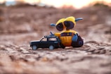 A yellow and blue toy robot with large eyes rests next to a small black toy car. They are placed on a sandy surface, with a blurred background suggesting a sunset or sunrise.