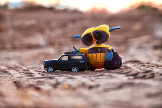 A yellow and blue toy robot with large eyes rests next to a small black toy car. They are placed on a sandy surface, with a blurred background suggesting a sunset or sunrise.