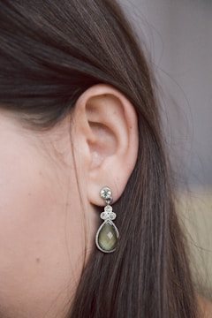 A close-up view of a woman's ear showcasing a dangling earring. The earring features a teardrop-shaped green stone with intricate silver detailing and small crystal embellishments above it. The background is softly blurred, highlighting the earring and the smooth, straight brown hair of the individual.