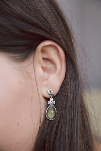 A close-up view of a woman's ear showcasing a dangling earring. The earring features a teardrop-shaped green stone with intricate silver detailing and small crystal embellishments above it. The background is softly blurred, highlighting the earring and the smooth, straight brown hair of the individual.