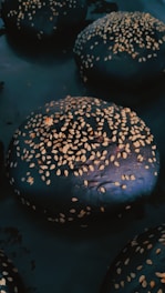 Several dark burger buns topped with sesame seeds are arranged in close proximity. The buns have a glossy finish and are set against a dark background.