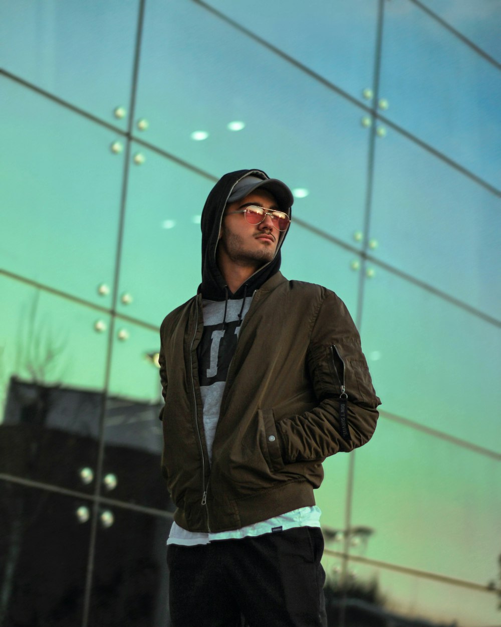 man wearing gray zip-up jacket and sunglasses standing while glancing his left side near glass walled building
