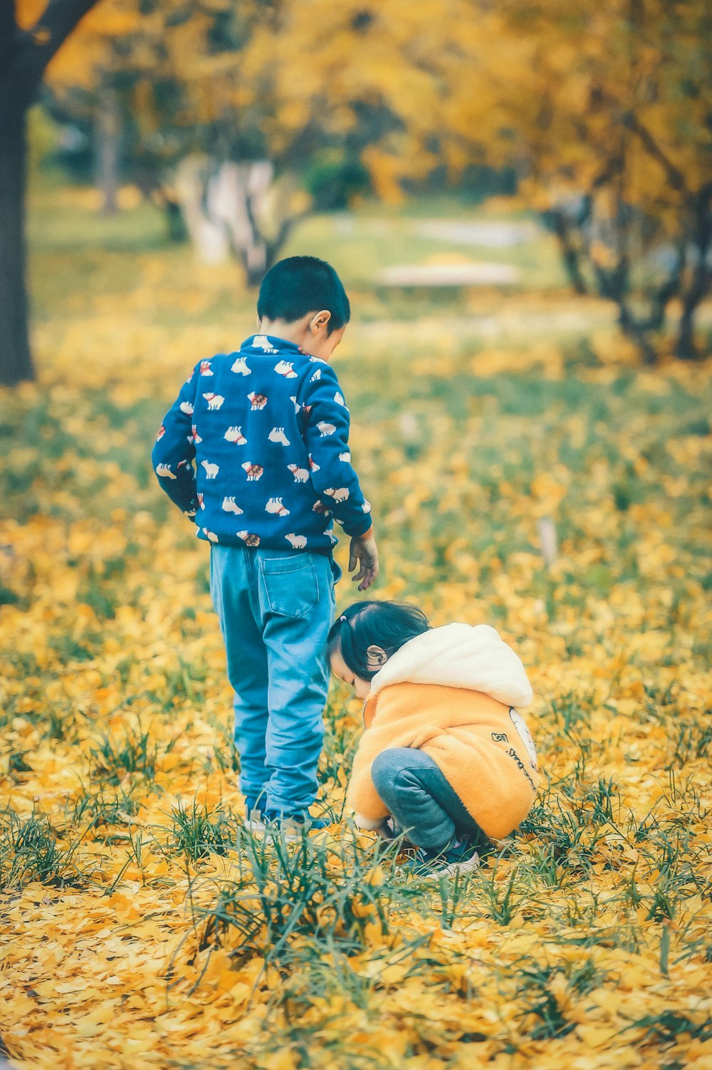 child wearing orange and white jacket picking grass and another child standing while looking down