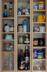 A wooden cabinet with multiple shelves is filled with various food items, jars, and bottles. The top shelves contain cooking oils, spices, and dry goods. There are jars of bouillon, honey, nuts, and spreads, along with glassware and mugs of different designs. On the lower shelves, there are larger bottles of liquor and a few empty glass containers.
