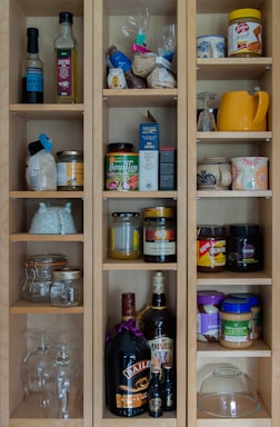 A wooden cabinet with multiple shelves is filled with various food items, jars, and bottles. The top shelves contain cooking oils, spices, and dry goods. There are jars of bouillon, honey, nuts, and spreads, along with glassware and mugs of different designs. On the lower shelves, there are larger bottles of liquor and a few empty glass containers.