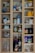 A wooden cabinet with multiple shelves is filled with various food items, jars, and bottles. The top shelves contain cooking oils, spices, and dry goods. There are jars of bouillon, honey, nuts, and spreads, along with glassware and mugs of different designs. On the lower shelves, there are larger bottles of liquor and a few empty glass containers.