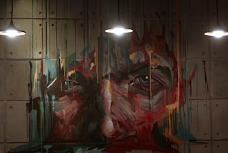 An abstract mural painting of a face is depicted with vivid brush strokes and dripping paint on a concrete wall. The art is illuminated by three overhead lights, enhancing the texture and colors.