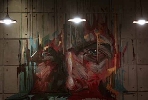 An abstract mural painting of a face is depicted with vivid brush strokes and dripping paint on a concrete wall. The art is illuminated by three overhead lights, enhancing the texture and colors.