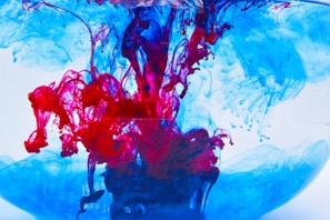 Vibrant swirls of red and blue ink are suspended in clear water. The colors intertwine and diffuse, creating intricate, fluid patterns that resemble abstract art. The contrast between the bright red and cool blue creates a dynamic visual effect.