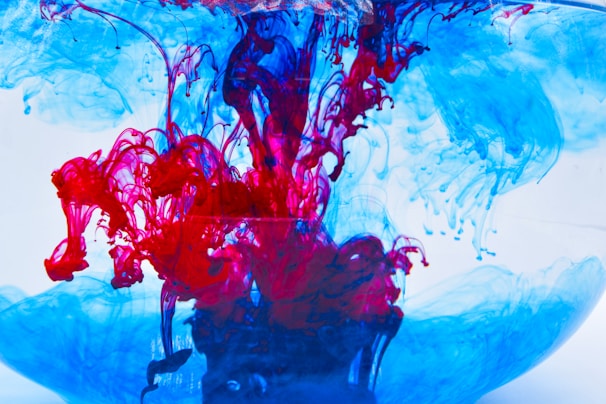 Vibrant swirls of red and blue ink are suspended in clear water. The colors intertwine and diffuse, creating intricate, fluid patterns that resemble abstract art. The contrast between the bright red and cool blue creates a dynamic visual effect.