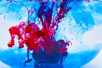 Vibrant swirls of red and blue ink are suspended in clear water. The colors intertwine and diffuse, creating intricate, fluid patterns that resemble abstract art. The contrast between the bright red and cool blue creates a dynamic visual effect.