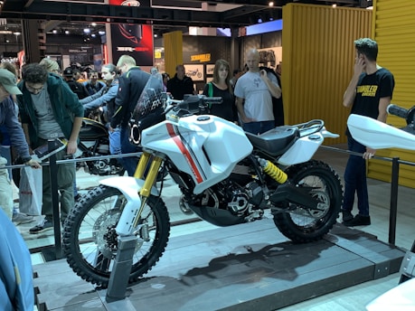A motorcycle is displayed on a platform in what appears to be an exhibition or showroom. The motorcycle is predominantly white with red and black accents, featuring a rugged design with knobby tires. Multiple people are gathered around, examining the motorcycle with interest. The setting includes various branded displays and posters in the background.