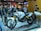 A motorcycle is displayed on a platform in what appears to be an exhibition or showroom. The motorcycle is predominantly white with red and black accents, featuring a rugged design with knobby tires. Multiple people are gathered around, examining the motorcycle with interest. The setting includes various branded displays and posters in the background.