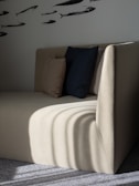 A minimalist beige couch with two cushions, one dark blue and one light brown, placed against a wall. The wall features a pattern of fish silhouettes, creating an artistic backdrop. The scene is partially illuminated, casting shadows on the couch and floor.