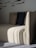 A minimalist beige couch with two cushions, one dark blue and one light brown, placed against a wall. The wall features a pattern of fish silhouettes, creating an artistic backdrop. The scene is partially illuminated, casting shadows on the couch and floor.
