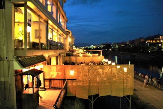 A lively evening scene featuring a well-lit restaurant with outdoor seating along a riverbank. Patrons are visible through screens, enjoying their meals under warm light. The background includes a dark blue sky, city lights, and a walkway beside the river with people strolling.