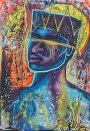 A vibrant street art mural featuring a face with a prominent crown, characterized by bold colors and dynamic brush strokes. The background includes swirling graffiti elements and splatters, creating a sense of movement and energy.