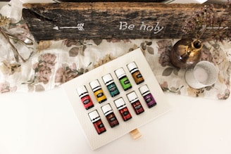 A set of essential oil bottles arranged on a textured white surface. The bottles are colorful, each labeled with a different name. Surrounding the bottles is a rustic, wooden sign with an arrow and the words 'Be holy' inscribed. A glass vase with branches, a metallic vase with dried flowers, and a decorative candle holder are also present on a floral-patterned cloth.