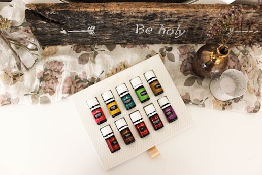A set of essential oil bottles arranged on a textured white surface. The bottles are colorful, each labeled with a different name. Surrounding the bottles is a rustic, wooden sign with an arrow and the words 'Be holy' inscribed. A glass vase with branches, a metallic vase with dried flowers, and a decorative candle holder are also present on a floral-patterned cloth.