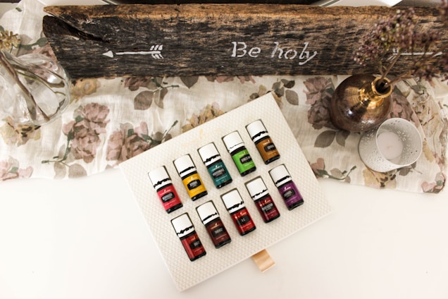 A set of essential oil bottles arranged on a textured white surface. The bottles are colorful, each labeled with a different name. Surrounding the bottles is a rustic, wooden sign with an arrow and the words 'Be holy' inscribed. A glass vase with branches, a metallic vase with dried flowers, and a decorative candle holder are also present on a floral-patterned cloth.