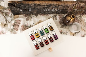 A set of essential oil bottles arranged on a textured white surface. The bottles are colorful, each labeled with a different name. Surrounding the bottles is a rustic, wooden sign with an arrow and the words 'Be holy' inscribed. A glass vase with branches, a metallic vase with dried flowers, and a decorative candle holder are also present on a floral-patterned cloth.