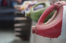 A row of colorful, weathered plastic jugs with handles is lined up along a concrete surface. The jugs have various colors such as red, green, and some mixed hues. The perspective is focused on the red jug in the foreground, with the background jugs becoming increasingly blurred.