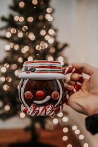 A hand is holding a festive ceramic mug shaped like a smiling snowman with red and white stripes. The background features a blurred Christmas tree adorned with soft, glowing lights.