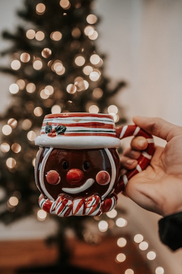 A hand is holding a festive ceramic mug shaped like a smiling snowman with red and white stripes. The background features a blurred Christmas tree adorned with soft, glowing lights.