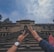 Two hands show a peace sign in the foreground, with an ornate, historical building in the background. The building features a large entrance and multiple arched windows. People are sitting on the steps leading up to the building. The sky is partly cloudy.