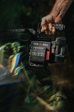 A hand grips a professional video camera, specifically a RED camera model, with a rugged design and various controls. The scene includes a prism-like light effect creating rainbow colors across the image.