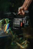 A hand grips a professional video camera, specifically a RED camera model, with a rugged design and various controls. The scene includes a prism-like light effect creating rainbow colors across the image.