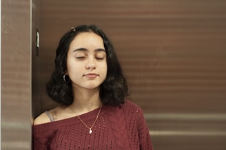 A young woman with short, dark hair stands against a wall with her eyes closed, wearing a maroon sweater and gold hoop earrings. She appears relaxed and calm.