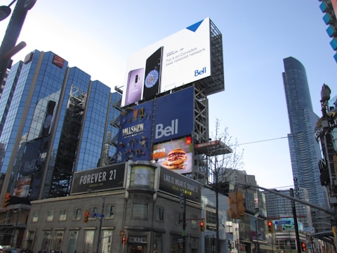 A bustling urban scene with tall buildings and numerous advertisements including prominent brands like Bell and McDonald's. The architecture features a mix of glass and steel structures, with large digital billboards displaying various products and services. There are traffic lights and street signs visible, contributing to the city's busy ambience.