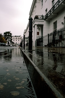 A rainy day scene features a wet sidewalk and street with visible puddles. The sidewalk is lined by a series of white, classical-style buildings with black wrought iron railings. Tall black posts are evenly spaced along the edge of the sidewalk. Raindrops create ripples in the water, and overcast skies add a dull, muted tone to the environment.