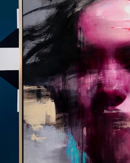 A close-up abstract painting featuring a face partially obscured by expressive brushstrokes. The colors are bold with a dominant pink hue over the face, contrasting against dark, blurred strokes on one side. The artwork has a mix of geometric and organic forms, with some sections appearing more structured while others are fluid and dynamic.