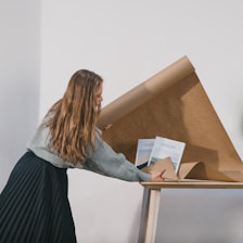 A person with long hair is unrolling brown kraft paper on a table, with two books visible underneath. The setting appears to be indoors against a plain white wall, creating a simple and minimalist atmosphere.