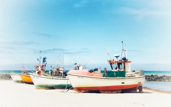 A group of fishing boats is resting on a sandy beach near a stone pier. The sea is calm and blue, with a clear sky above. The boats are colorful, mainly painted in shades of orange, red, and white, and appear to be well-equipped with radio antennas and other fishing gear.