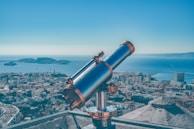 A telescope is positioned on a platform overlooking a coastal cityscape. The city is densely packed with buildings, and the sea stretches into the horizon with several small islands visible in the distance. The sky is clear, adding to the serene atmosphere.