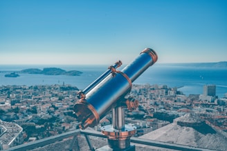 A telescope is positioned on a platform overlooking a coastal cityscape. The city is densely packed with buildings, and the sea stretches into the horizon with several small islands visible in the distance. The sky is clear, adding to the serene atmosphere.