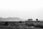 A vintage van is parked on a rural road amidst flat, barren fields, with mountainous terrain in the distant background. The van is loaded with luggage on the roof, suggesting a travel theme. The sky is overcast, adding a somber and contemplative mood to the scene.