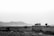 A vintage van is parked on a rural road amidst flat, barren fields, with mountainous terrain in the distant background. The van is loaded with luggage on the roof, suggesting a travel theme. The sky is overcast, adding a somber and contemplative mood to the scene.