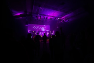 A dimly lit venue filled with silhouettes of people, directed towards a stage with vibrant purple lighting. A prominent screen displays the words 'OUR CITY'. The atmosphere suggests a concert or event as the participants appear engaged, some with raised hands.