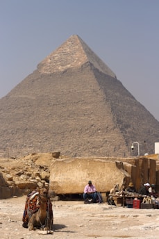 A large pyramid with a weathered surface dominates the background, set under a clear blue sky. In the foreground, a camel adorned with colorful blankets sits leisurely on the sandy ground. Nearby, there are several people seated, one of whom appears to be selling trinkets or souvenirs. The terrain is arid, with scattered stones and rock formations.