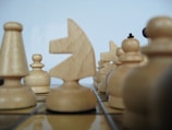Several wooden chess pieces are arranged on a chessboard. The focus is on the knight in the center, surrounded by pawns and rooks. The pieces have a natural wood grain texture and are positioned closely, suggesting a strategic game setup.