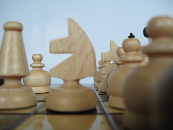 Several wooden chess pieces are arranged on a chessboard. The focus is on the knight in the center, surrounded by pawns and rooks. The pieces have a natural wood grain texture and are positioned closely, suggesting a strategic game setup.
