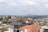 A sprawling residential area with a mix of modern and older architecture. Houses with red-tiled roofs are surrounded by greenery and scattered trees. In the background, rolling hills are visible under a partly cloudy sky. Power lines cross the image, adding an urban element.