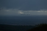 A dark, overcast landscape features a distant industrial facility emitting smoke, situated beneath heavy clouds. The foreground consists of shadowy silhouettes of trees or bushes, while the horizon is barely visible due to the atmospheric haze.