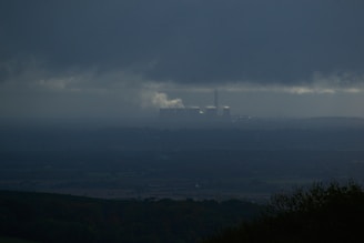 A dark, overcast landscape features a distant industrial facility emitting smoke, situated beneath heavy clouds. The foreground consists of shadowy silhouettes of trees or bushes, while the horizon is barely visible due to the atmospheric haze.